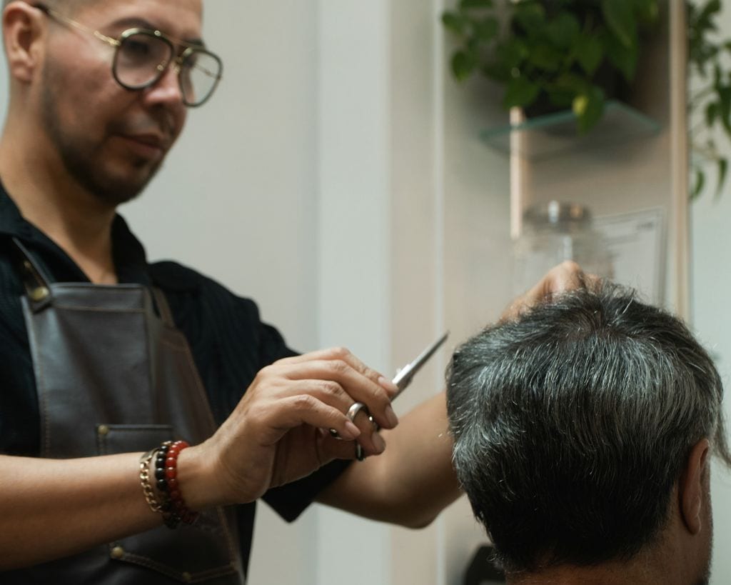 Moses cutting a client's hair with scissors, mirror and shelves behind